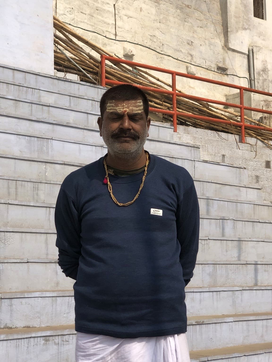 A man in a navy sweater and white dhoti stands on the ghats with religious markings on his forehead, Varanasi, 2019