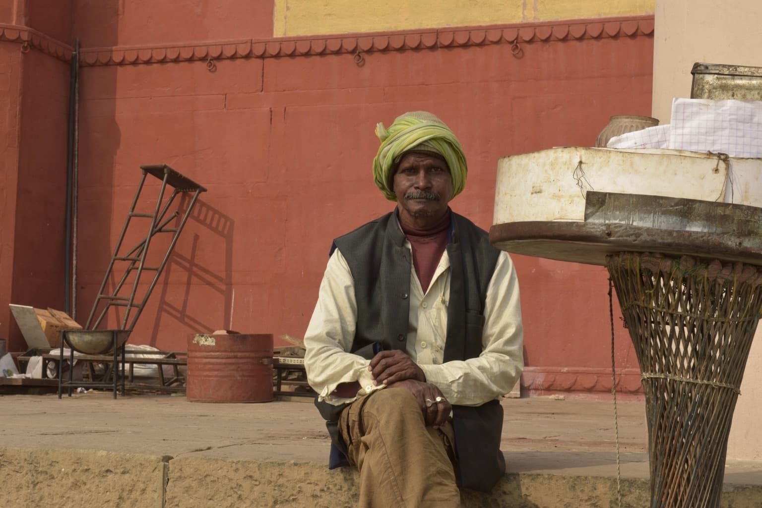A seated man in a green turban rests beside an elevated vendor tray on the ghats of Varanasi, 2019