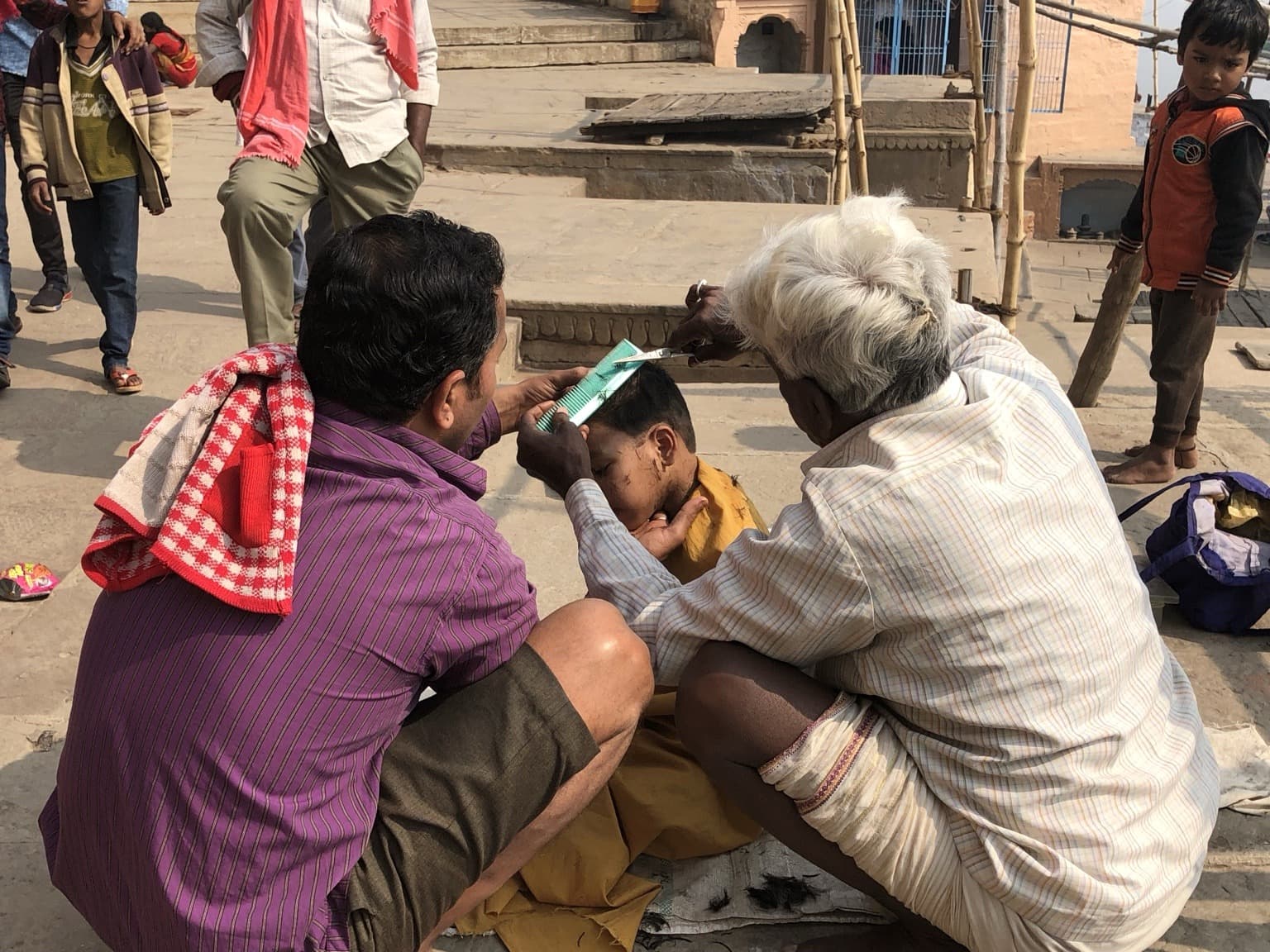 Two men cut a boy’s hair in a public space on the ghats of Varanasi, 2019