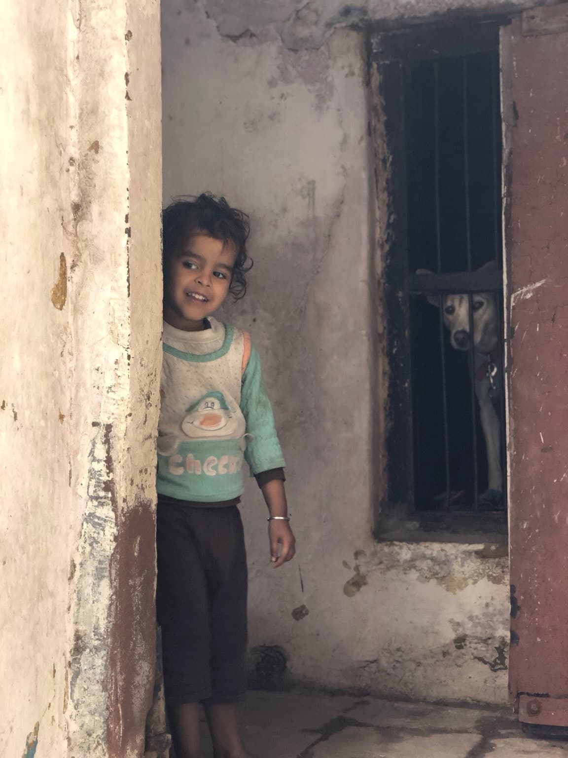 A child smiles from a doorway while a dog looks through bars in a nearby opening, Varanasi, 2019