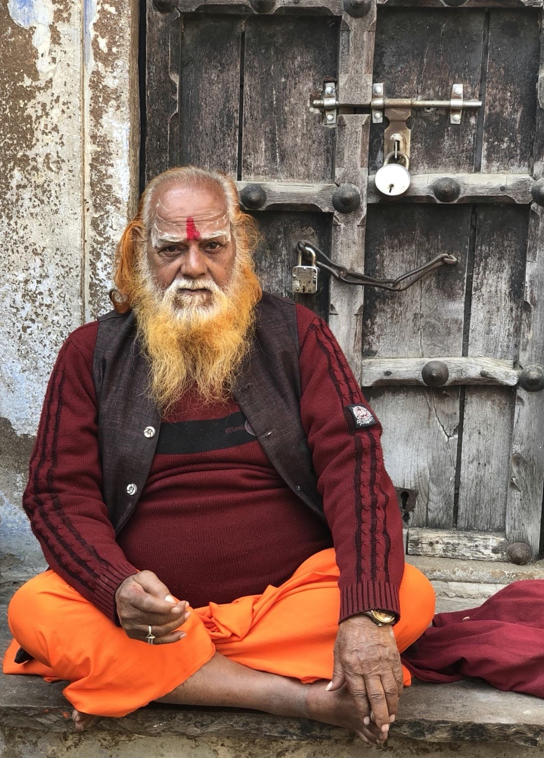 A seated holy man with an orange-dyed beard sits before a weathered locked wooden door in Varanasi, 2019