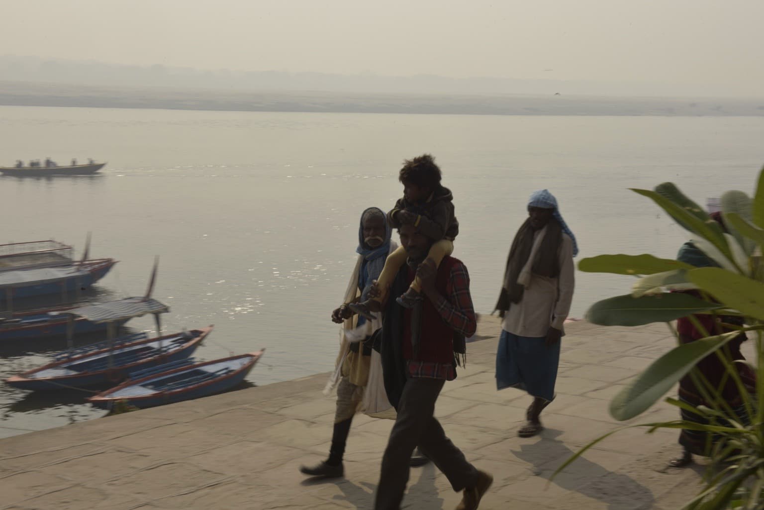 A man carries a child on his shoulders while walking beside the Ganges with boats behind him, Varanasi, 2019