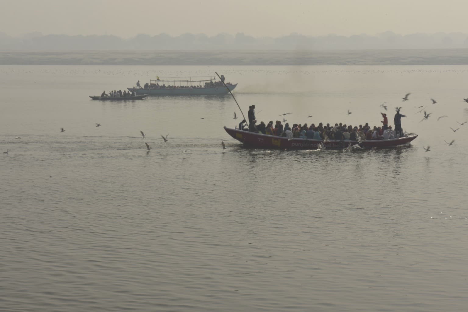 A crowded boat crosses the Ganges as birds fly close to the water in Varanasi, 2019