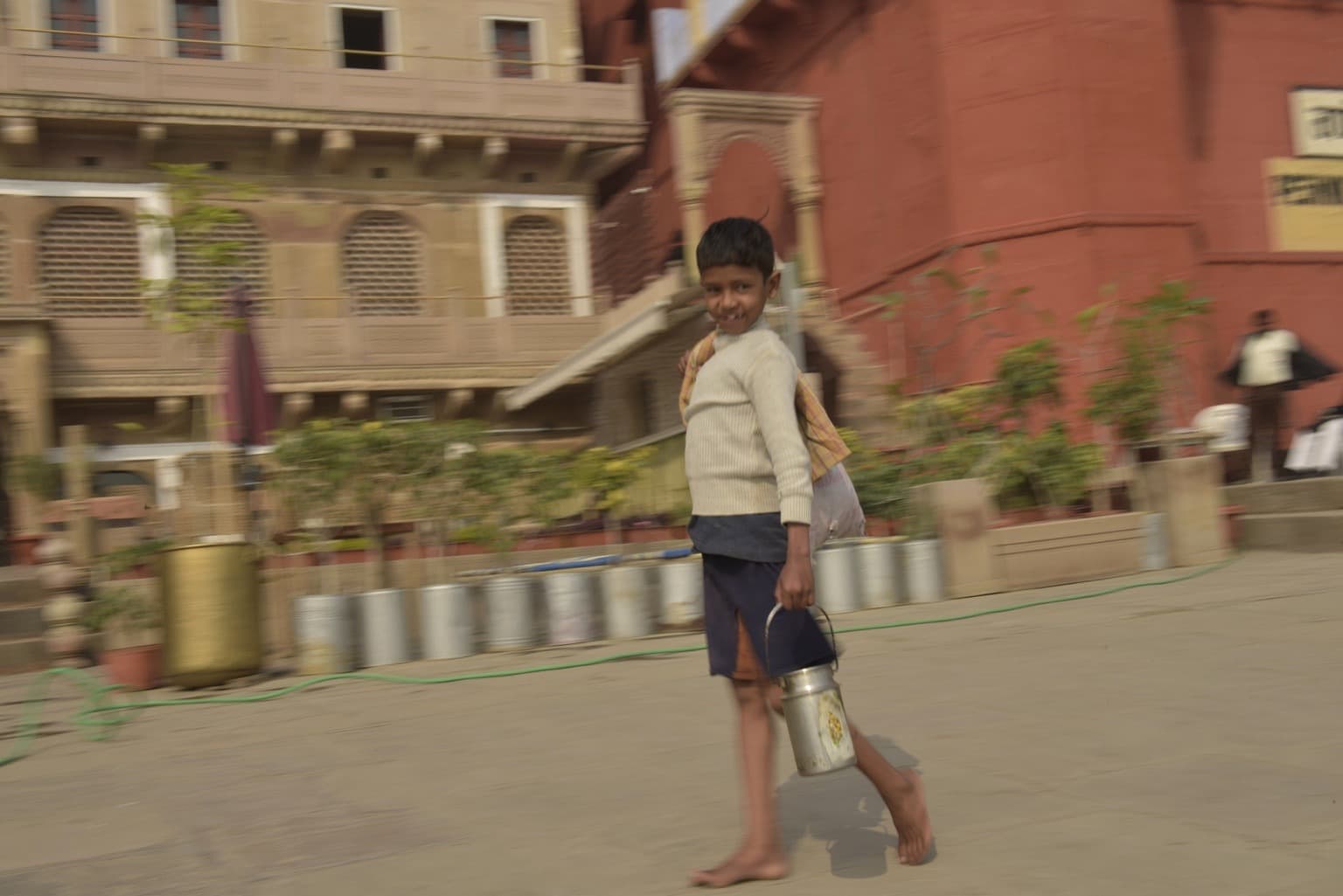 A barefoot boy carrying a small metal container walks across the ghats, captured with motion blur, Varanasi, 2019