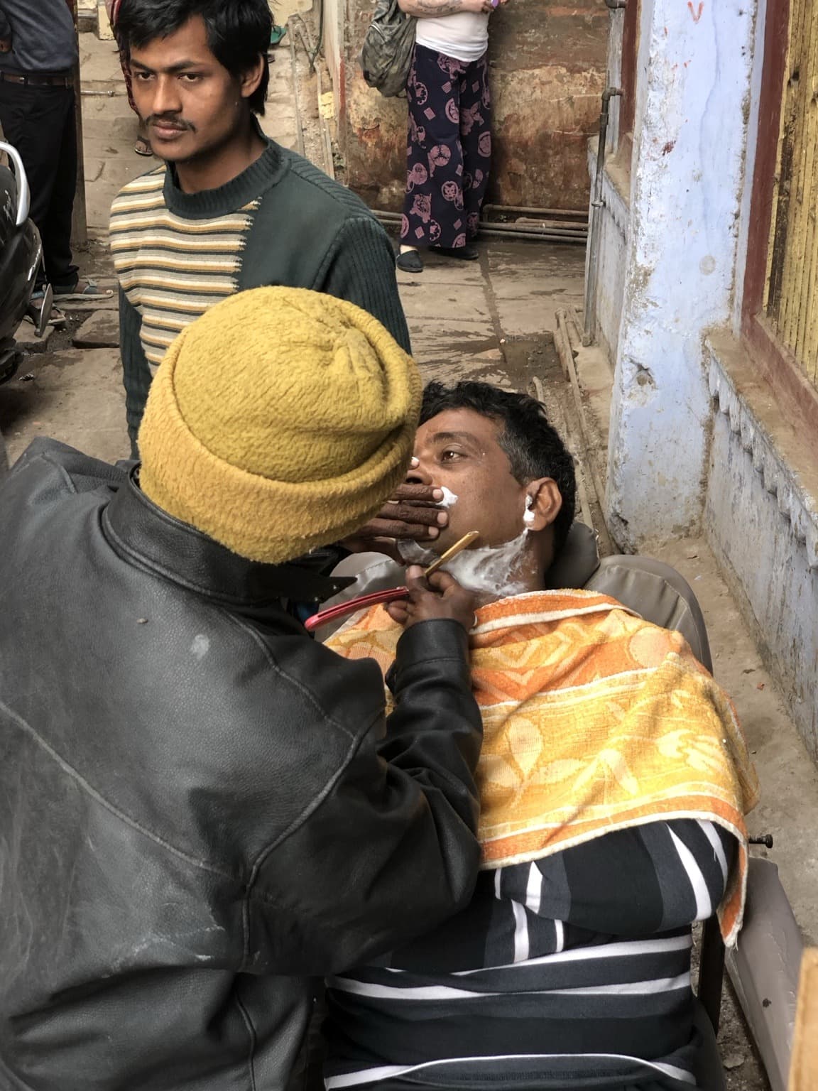 A barber shaves a reclining customer in a narrow outdoor lane in Varanasi, 2019