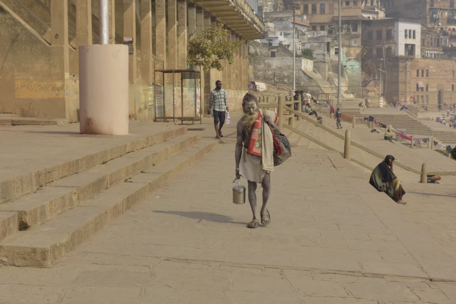 An ash-covered holy man carrying a small metal container walks along the ghats of Varanasi, 2019