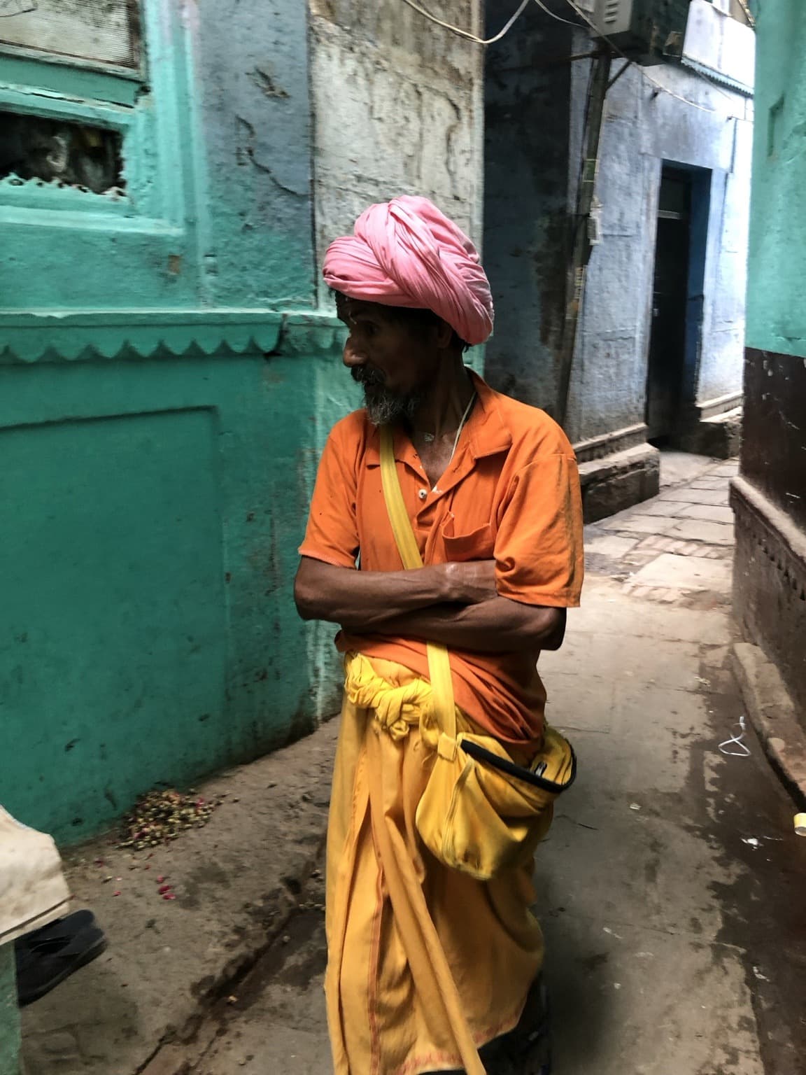 A closer portrait of a man in a pink turban, orange shirt and yellow cloth in a turquoise-painted lane in Varanasi, 2019