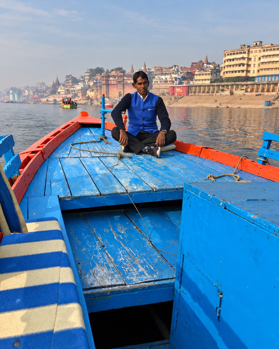 A boatman sits cross-legged on the blue deck of a wooden boat with the ghats receding behind him in morning haze, Varanasi, 2019