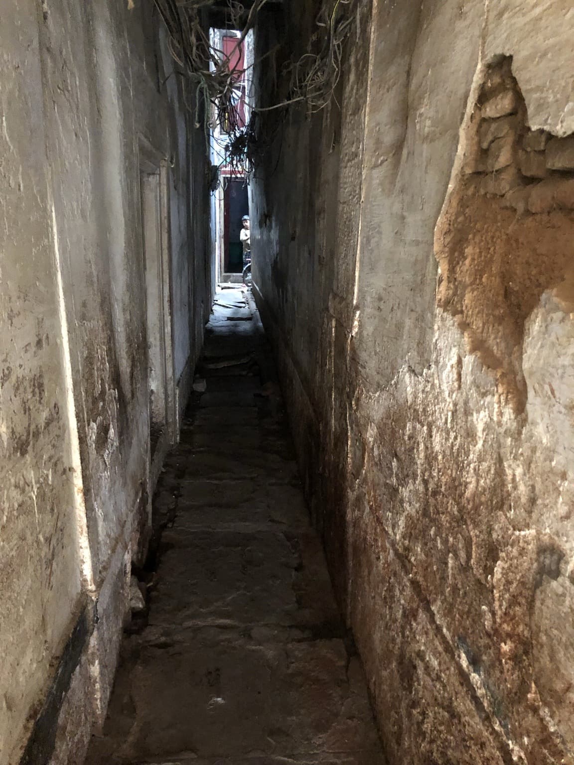A very narrow weathered lane recedes toward a small figure beneath tangled overhead wires in Varanasi, 2019