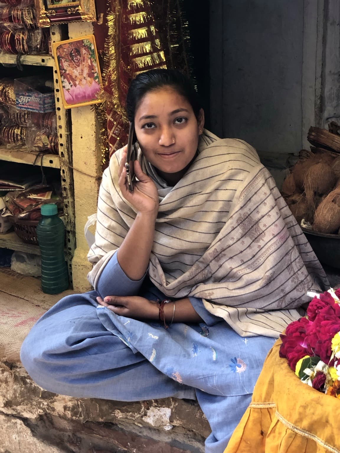A young woman sits at a small shop or stall speaking on a mobile phone, with flowers and goods nearby, Varanasi, 2019
