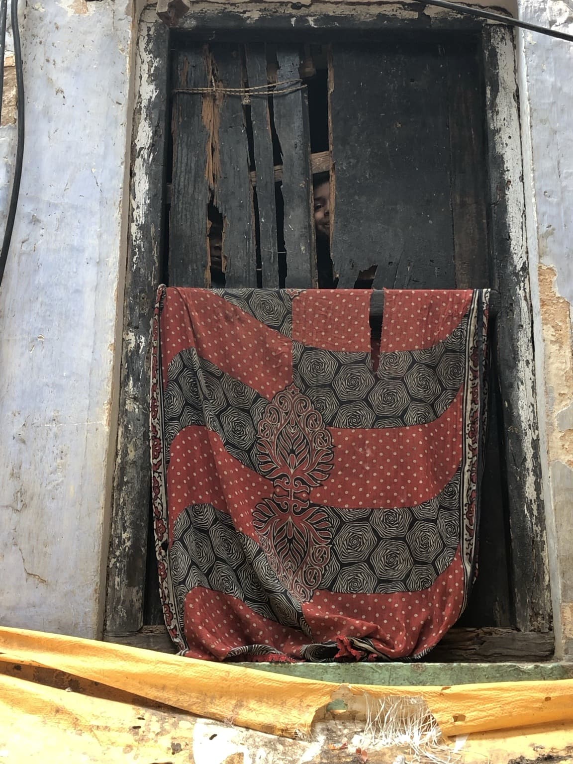 Children look through gaps in a broken wooden window above a patterned cloth in Varanasi, 2019