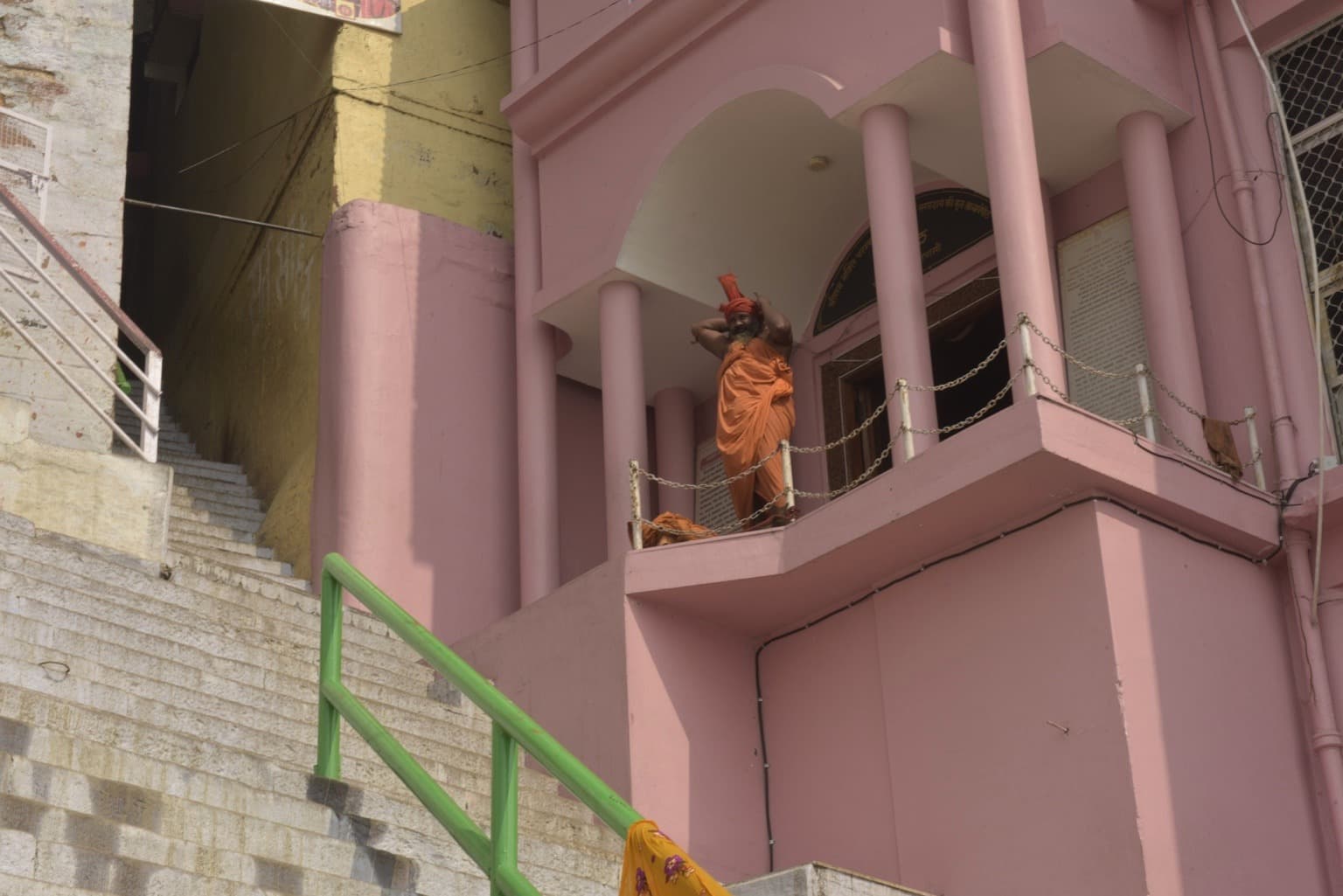 A woman in orange clothing stands on a balcony of a pink riverfront building above the stone steps in Varanasi, 2019
