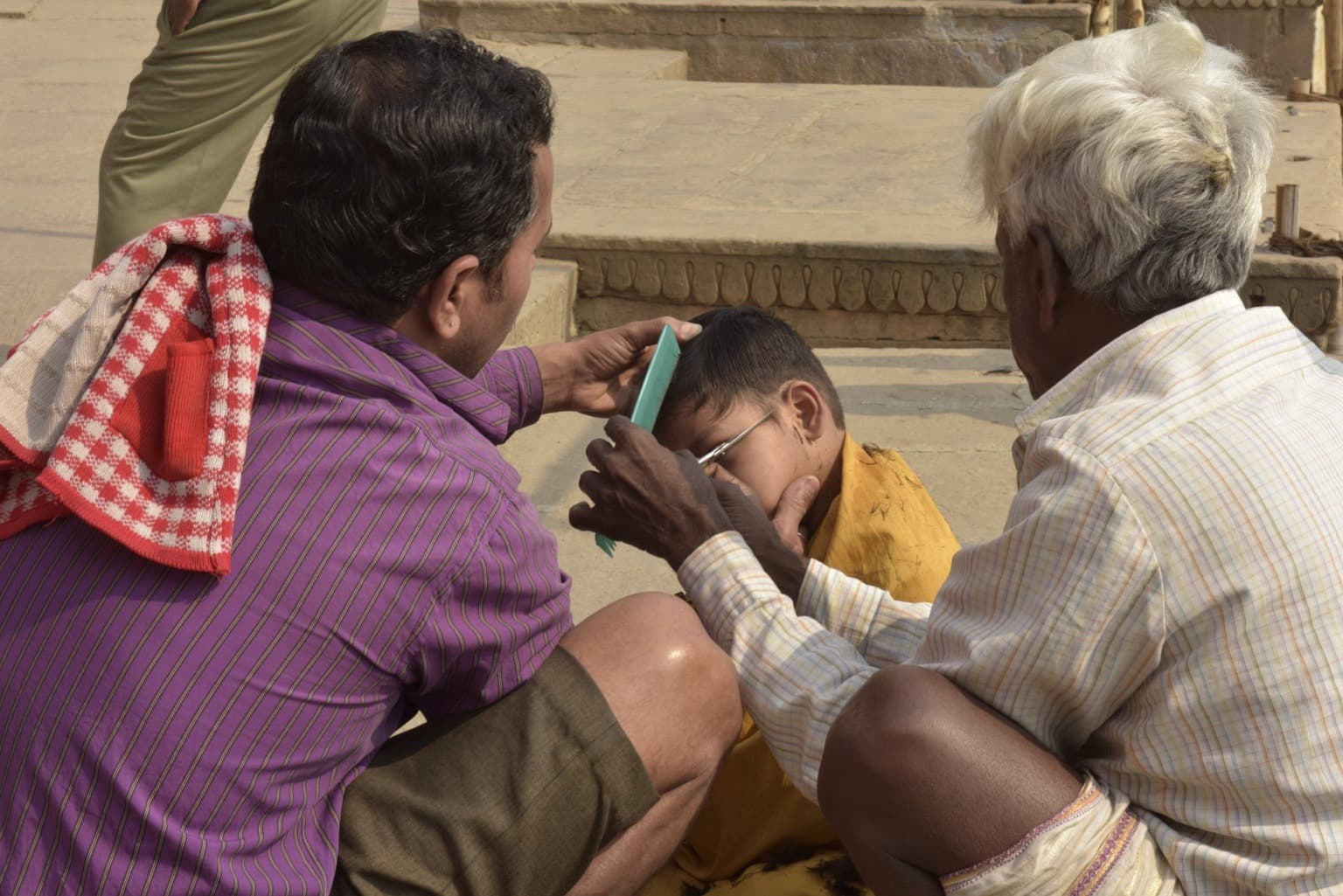 Two men carefully cut a boy’s hair on the ghats of Varanasi, 2019