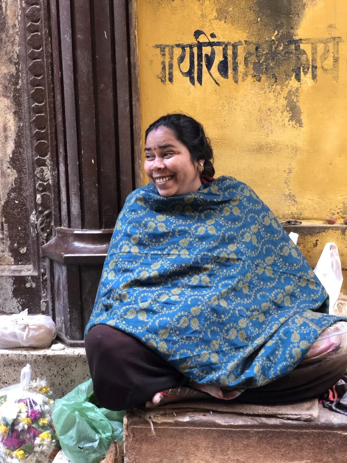 A smiling woman wrapped in a blue patterned shawl sits beside a yellow wall in Varanasi, 2019