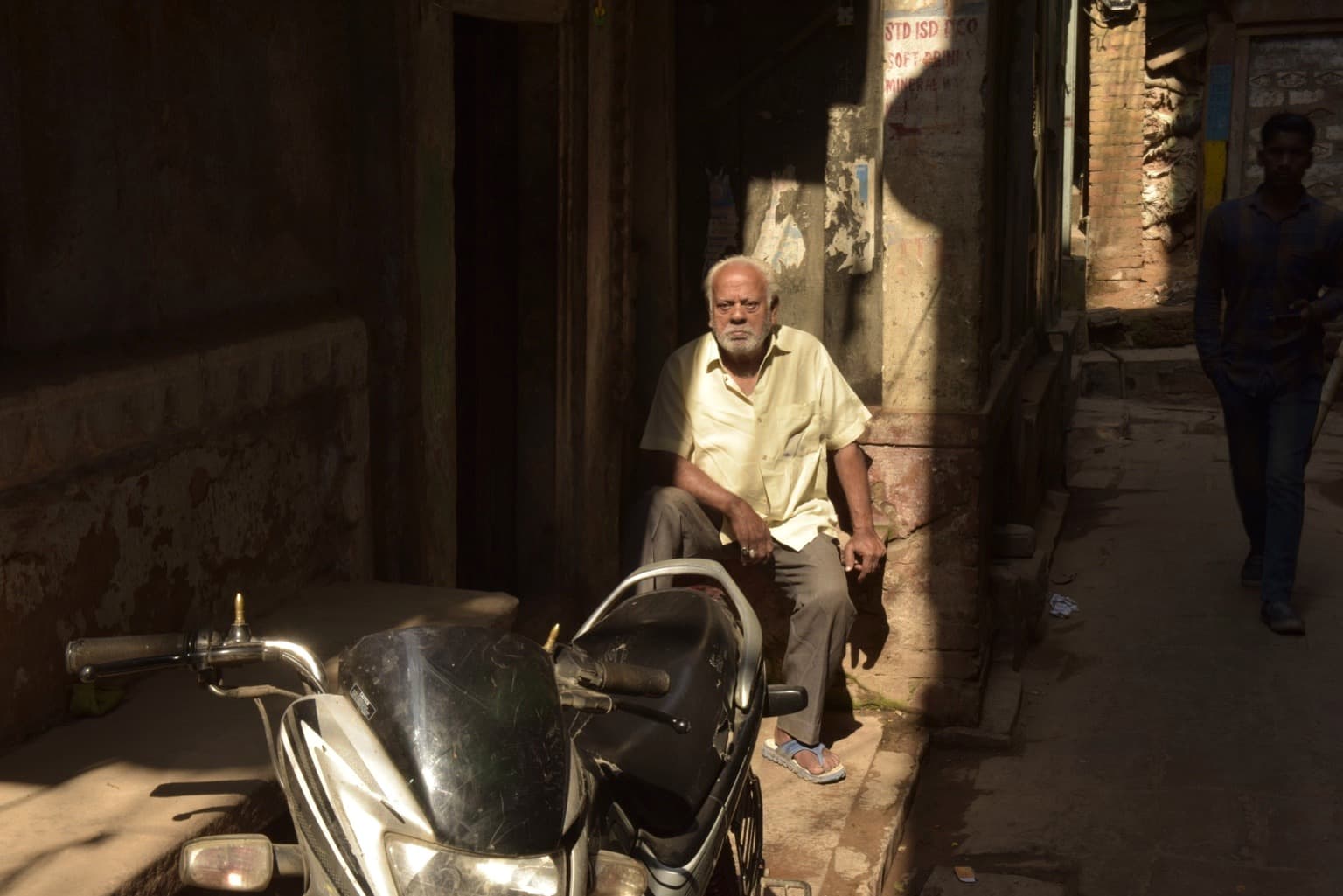 An older man sits in a narrow shaft of sunlight beside a parked motorcycle in a Varanasi lane, 2019