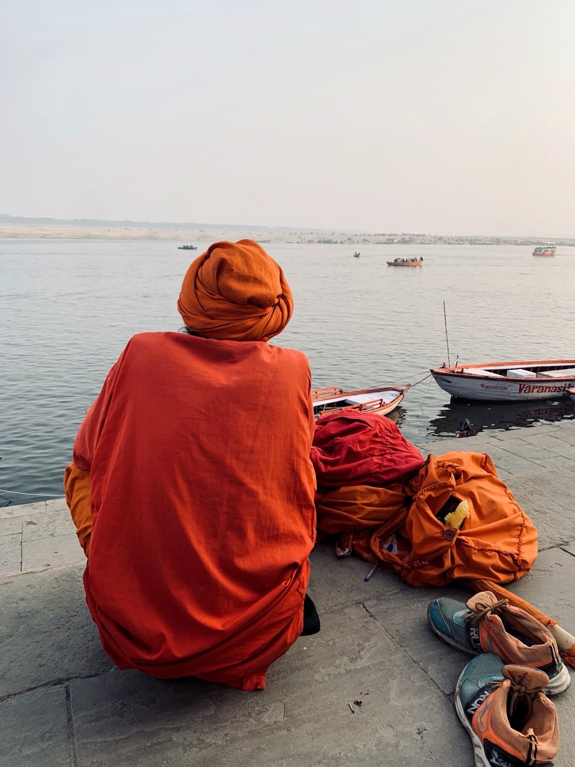 A turned figure is set against the broad Ganges, distant boats and haze in Varanasi, 2019.