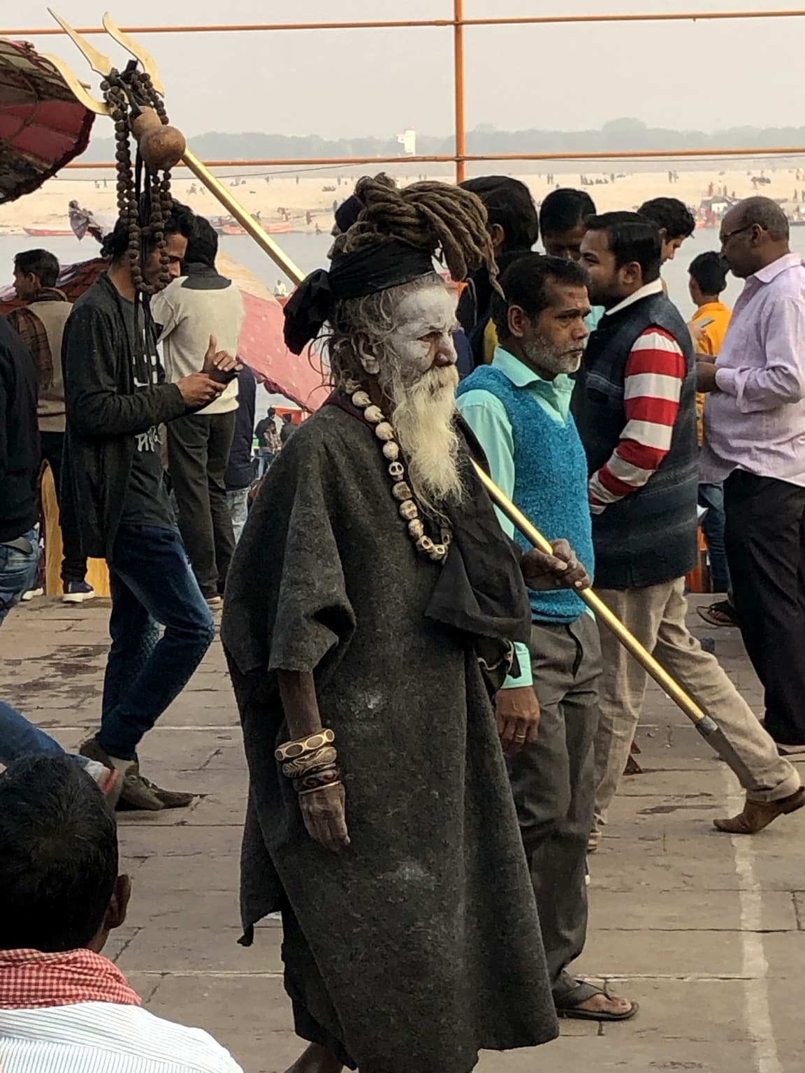 An ash-covered sadhu with matted hair and a long white beard carries a trident staff through the crowded ghats, with the Ganges behind him, Varanasi, 2019.