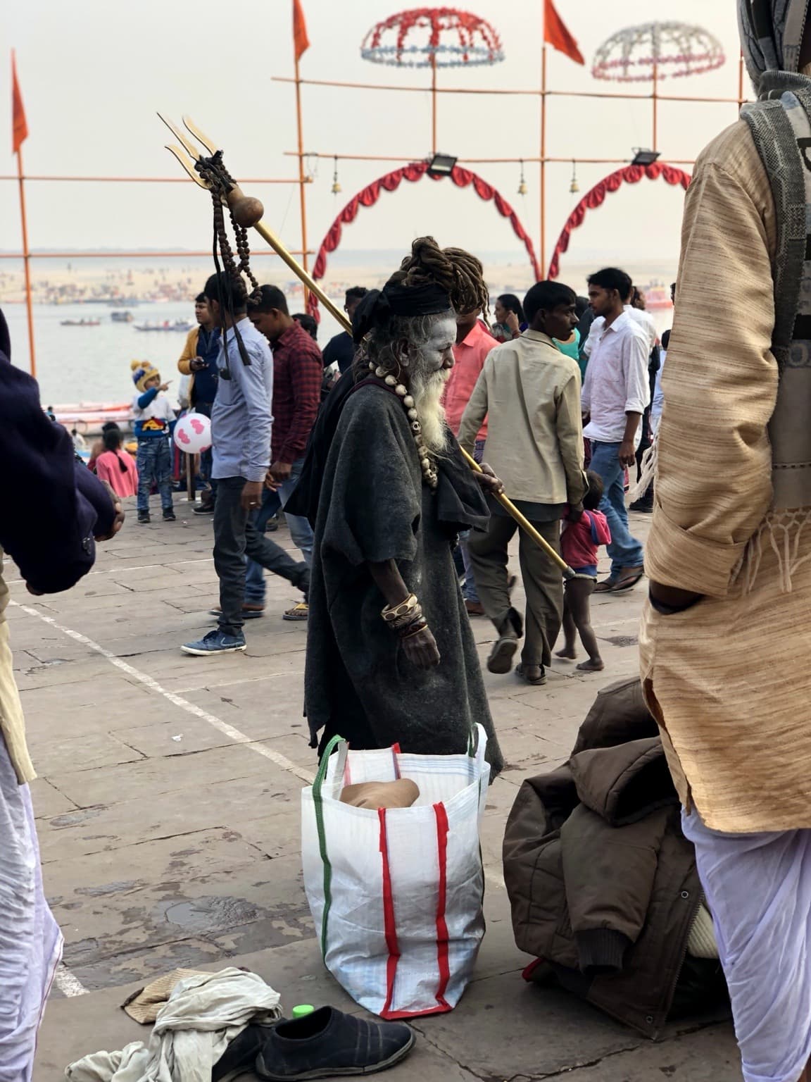 The same ash-covered sadhu stands on the ghats with his trident angled across the frame, surrounded by pedestrians and his belongings, Varanasi, 2019.