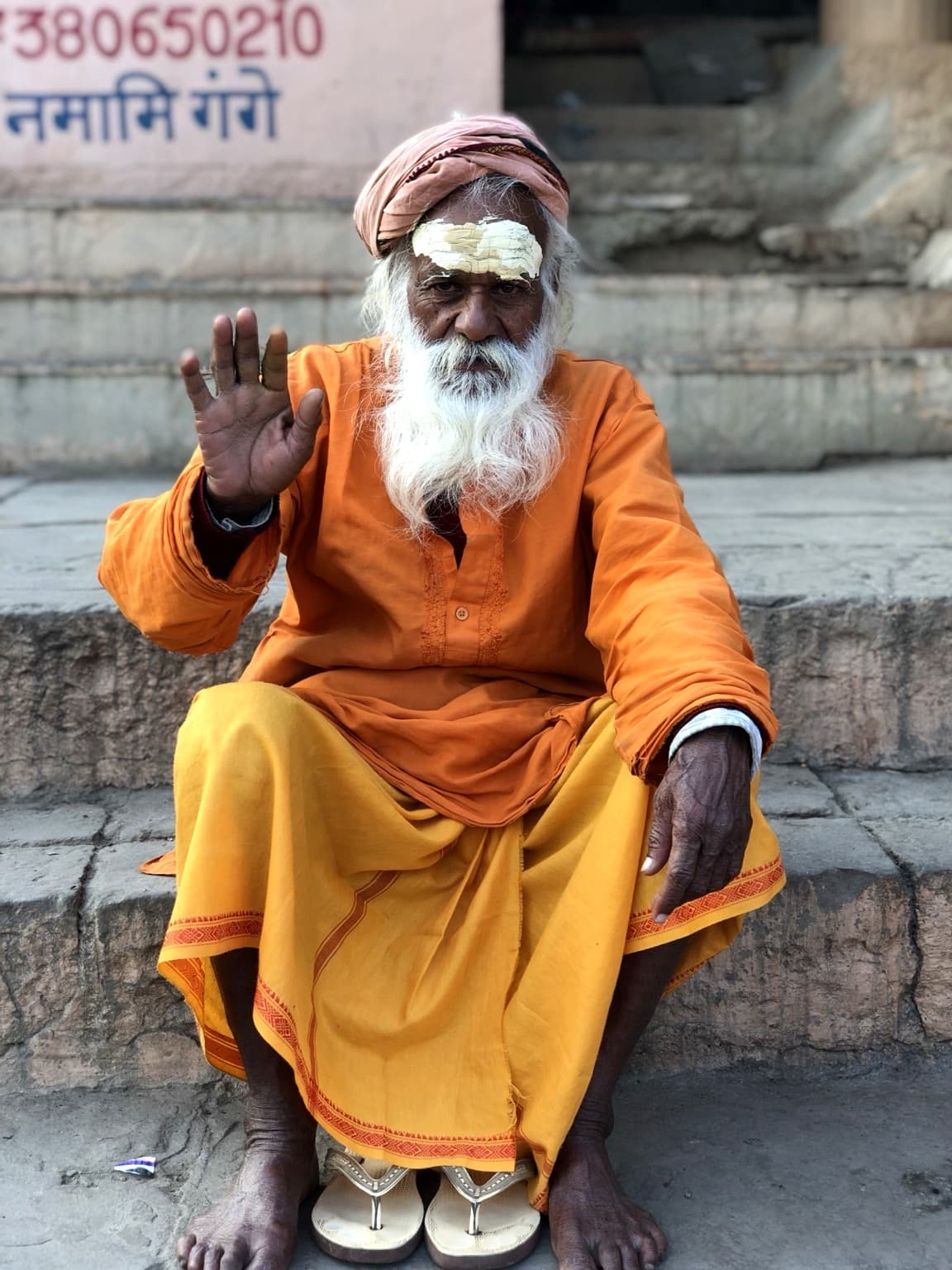 An elderly sadhu in orange and yellow robes sits on stone steps and raises one hand toward the camera, Varanasi, 2019.