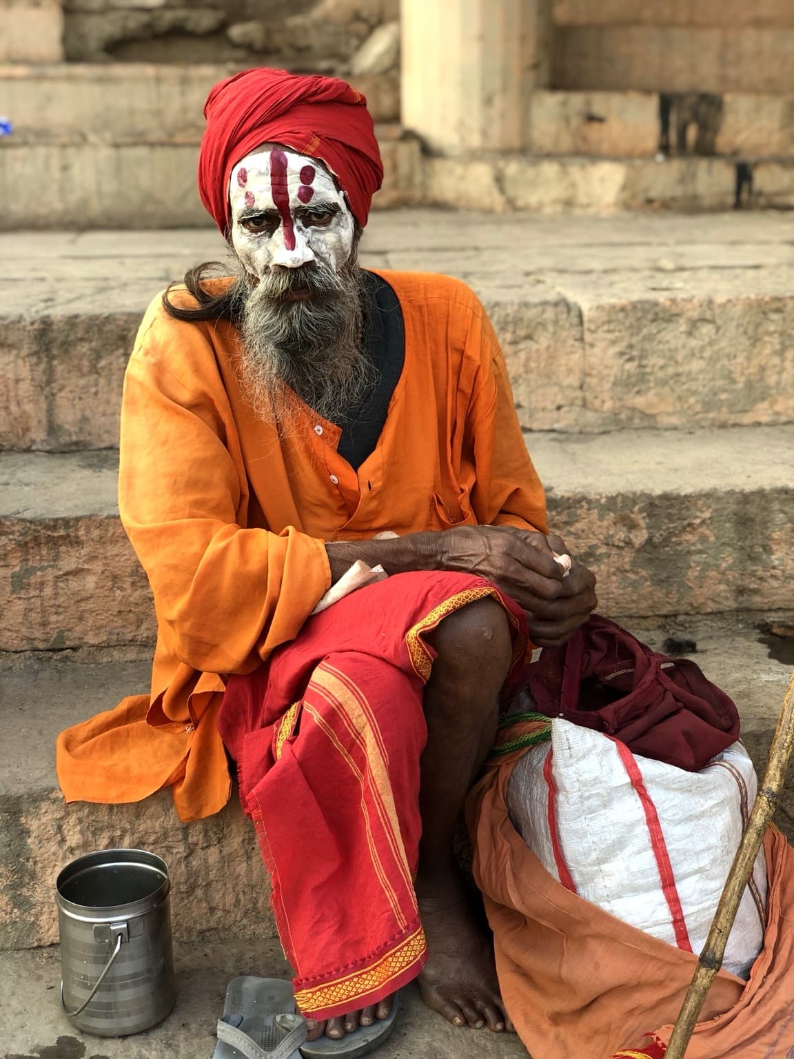 A seated sadhu in orange and red robes faces the camera with white and red markings painted across his face, Varanasi, 2019.