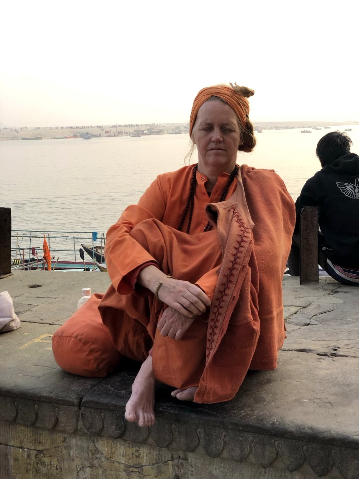 A woman in orange robes sits quietly beside the Ganges with her eyes closed, boats and the far bank behind her, Varanasi, 2019.