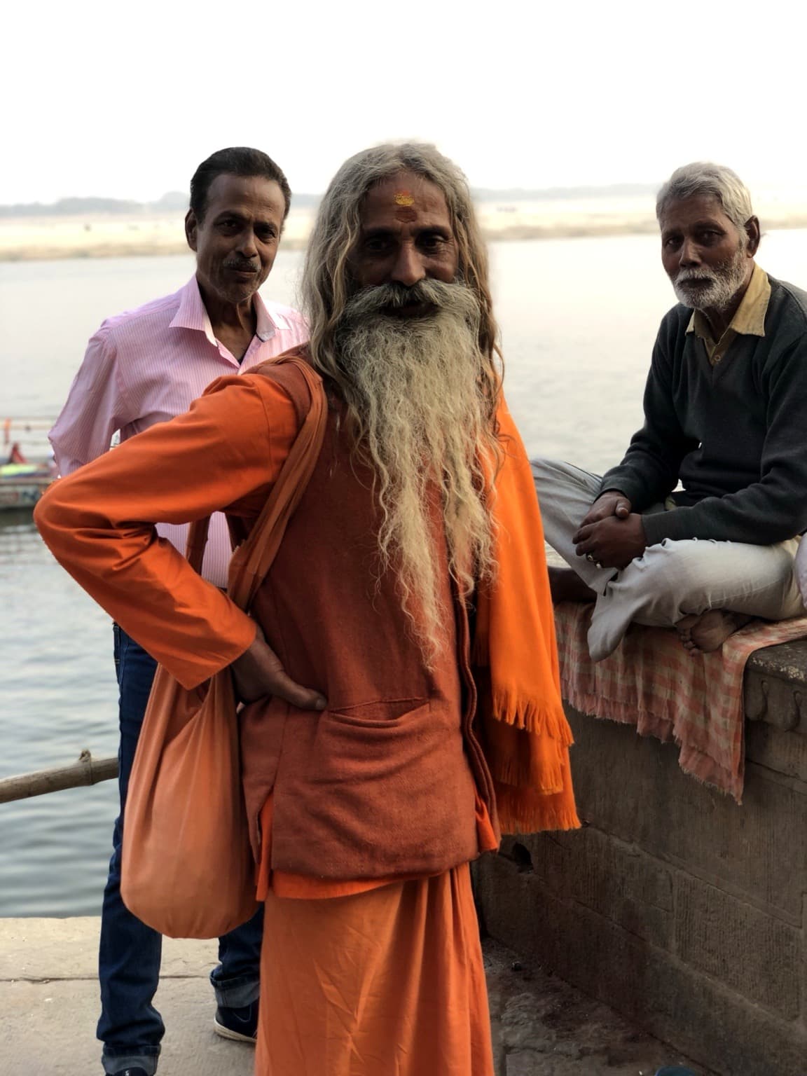 A long-haired, bearded sadhu in orange robes stands close to the Ganges while two men remain behind him, Varanasi, 2019.
