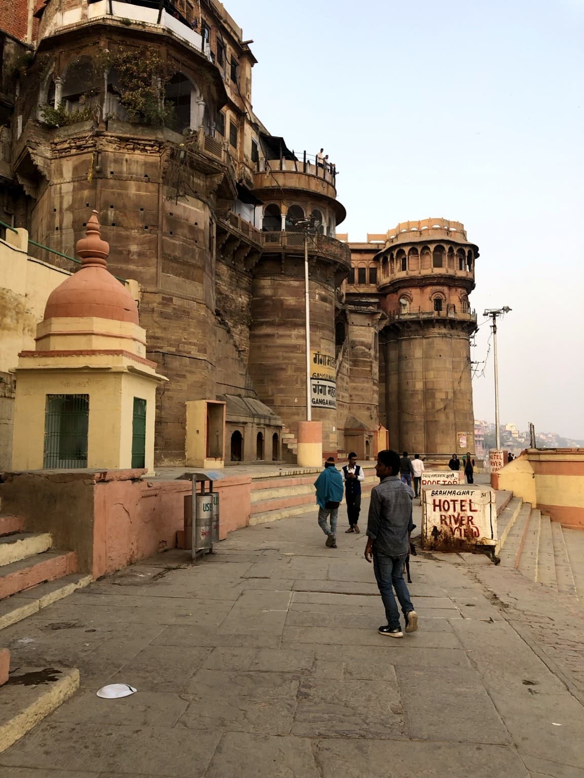 Pedestrians walk beside monumental riverfront architecture and stone steps on the ghats of Varanasi, 2019