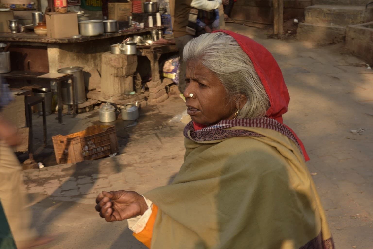 An older woman wrapped in a shawl moves through a ghat-side market area in warm light, Varanasi, 2019