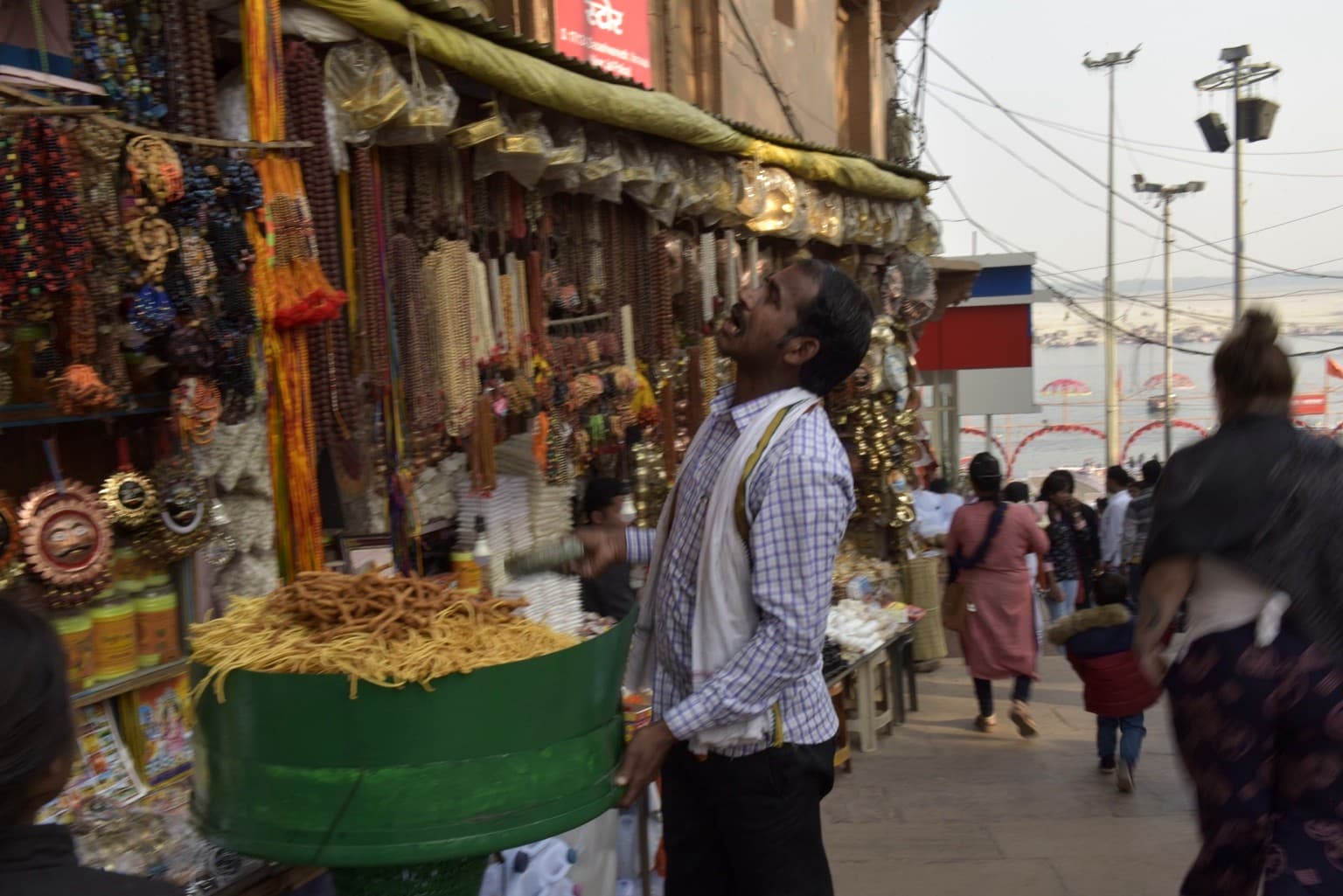 A man looks upward at hanging goods at a densely stocked stall, with the Ganges visible beyond the steps, Varanasi, 2019