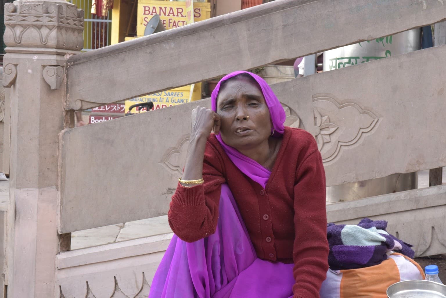 A woman in a vivid magenta sari and red sweater sits beside her belongings near the ghat steps, Varanasi, 2019