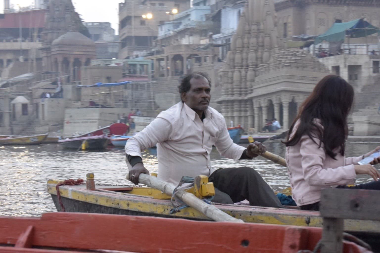 A boatman rows a passenger boat on the Ganges with riverfront temples and moored boats behind him, Varanasi, 2019
