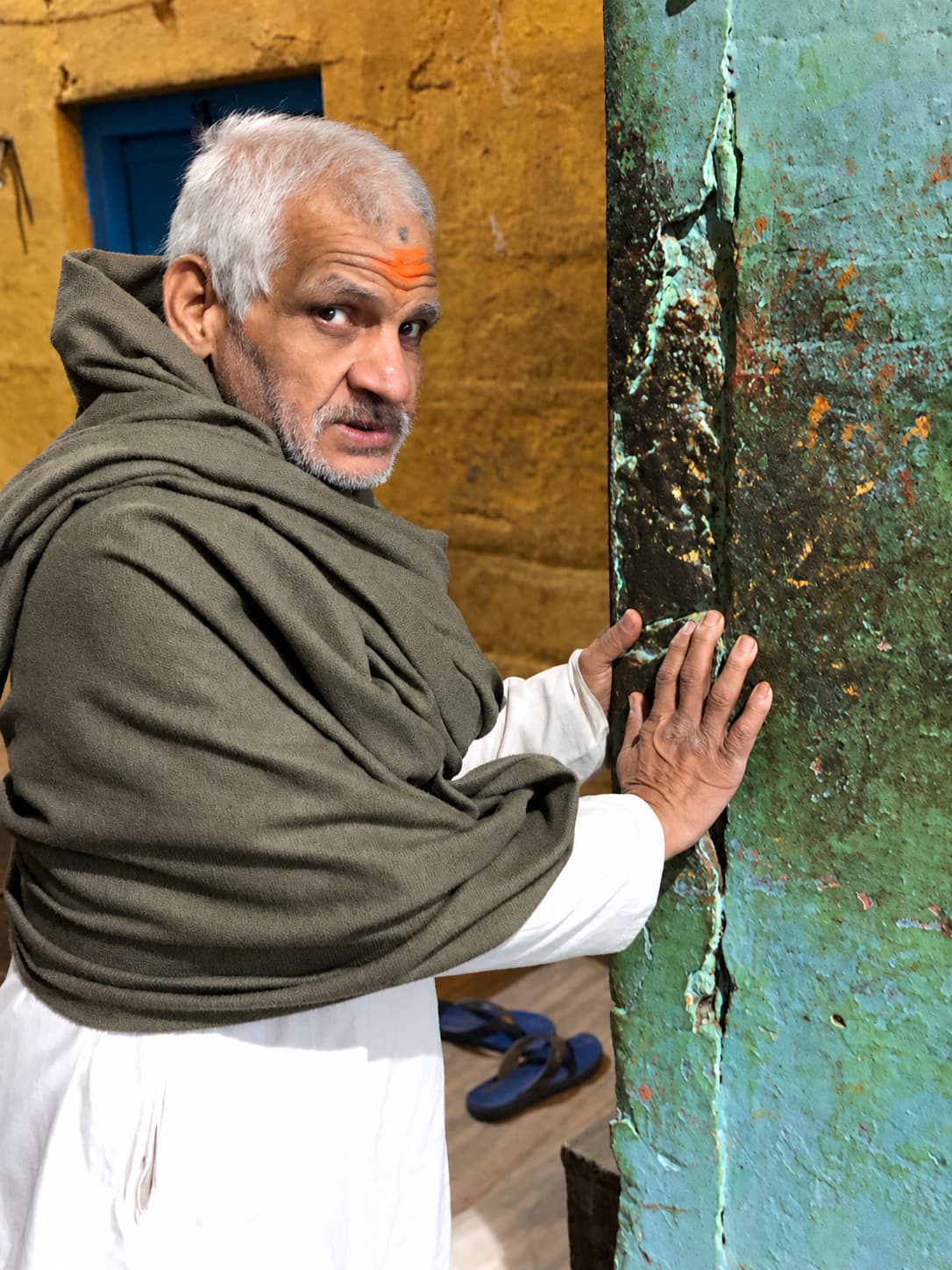 An older man wrapped in an olive shawl looks toward the camera while resting both hands against a weathered green-painted wall, Varanasi, 2019