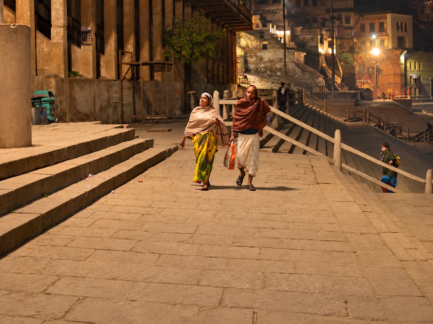 A woman and man walk together along a broad stone ghat at night while a child moves beside the railing, Varanasi, 2019
