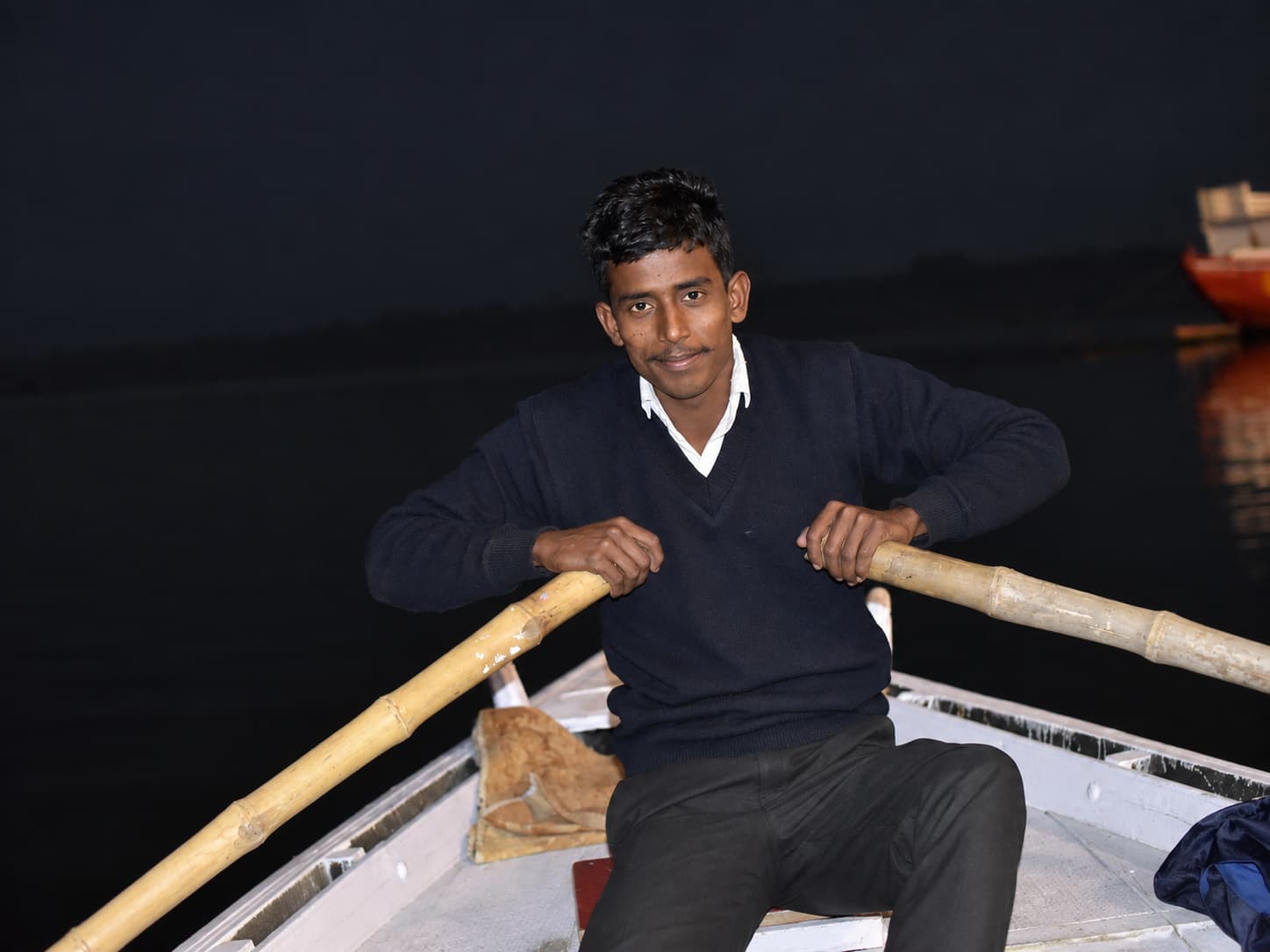 A young boatman sits in a small rowboat at night and looks directly toward the camera with both hands resting on the oars, Varanasi, 2019