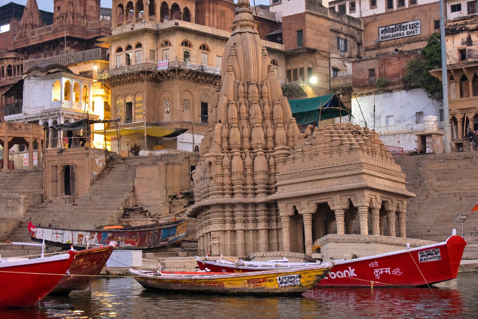 A stone temple tower rises above moored red boats along the Ganges in Varanasi, 2019