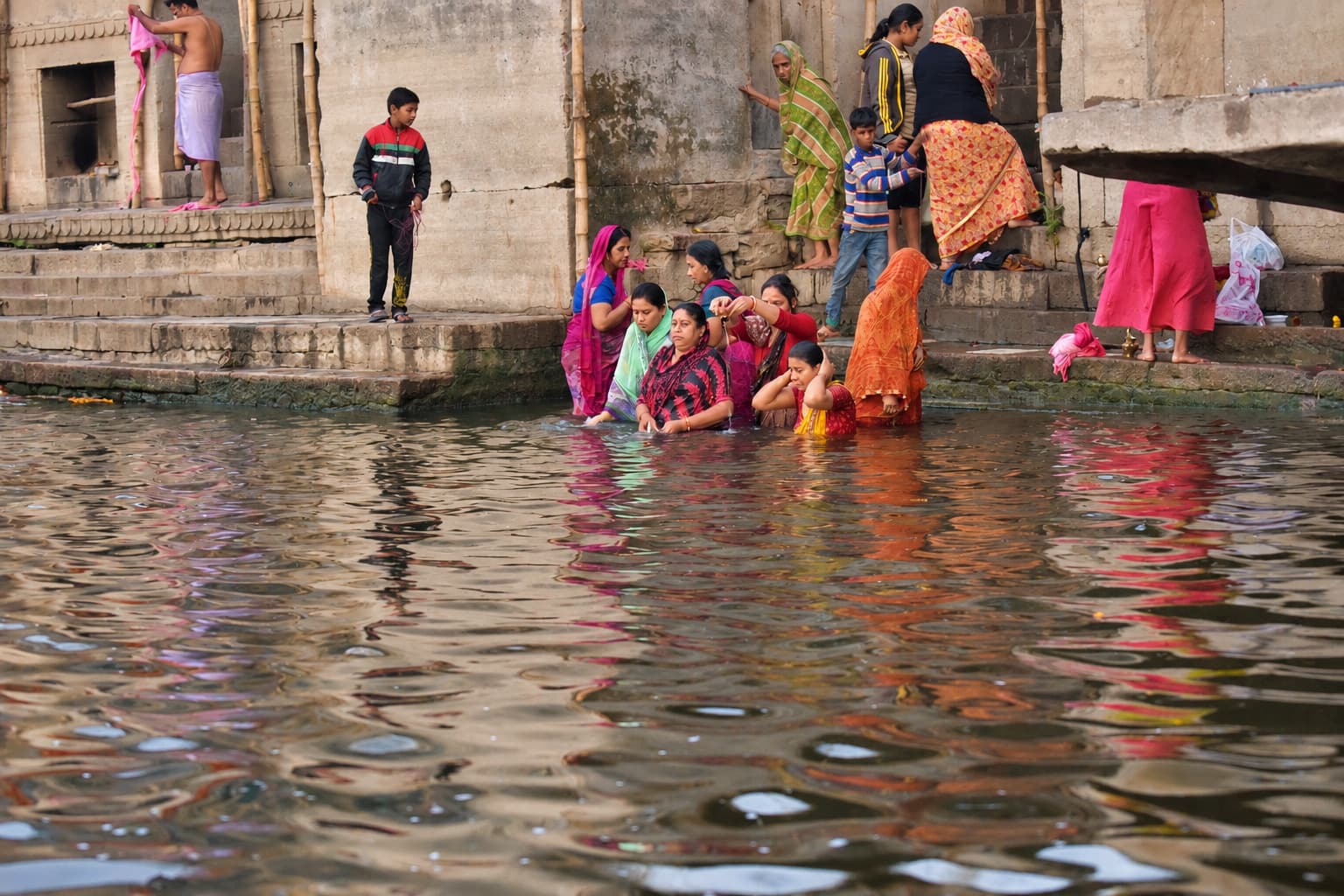 Women in colourful clothing bathe and wash at the edge of the Ganges below stone steps in Varanasi, 2019