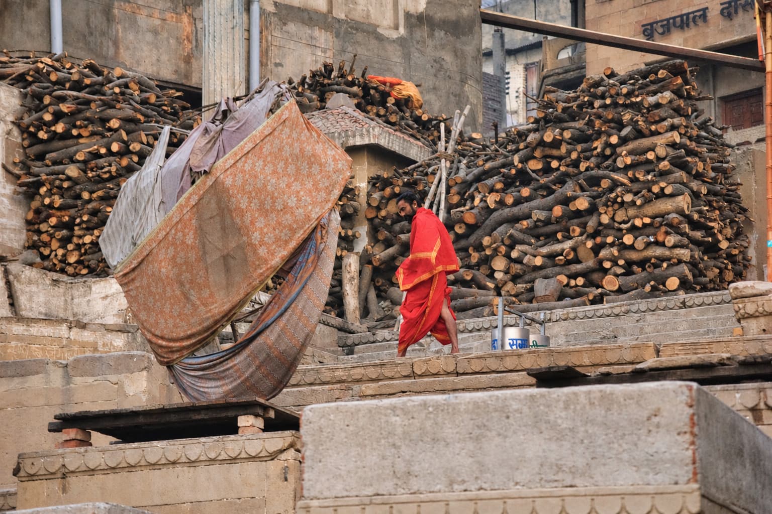 Large stacks of firewood rise beside a patterned cloth and stone structures on the ghats of Varanasi, 2019