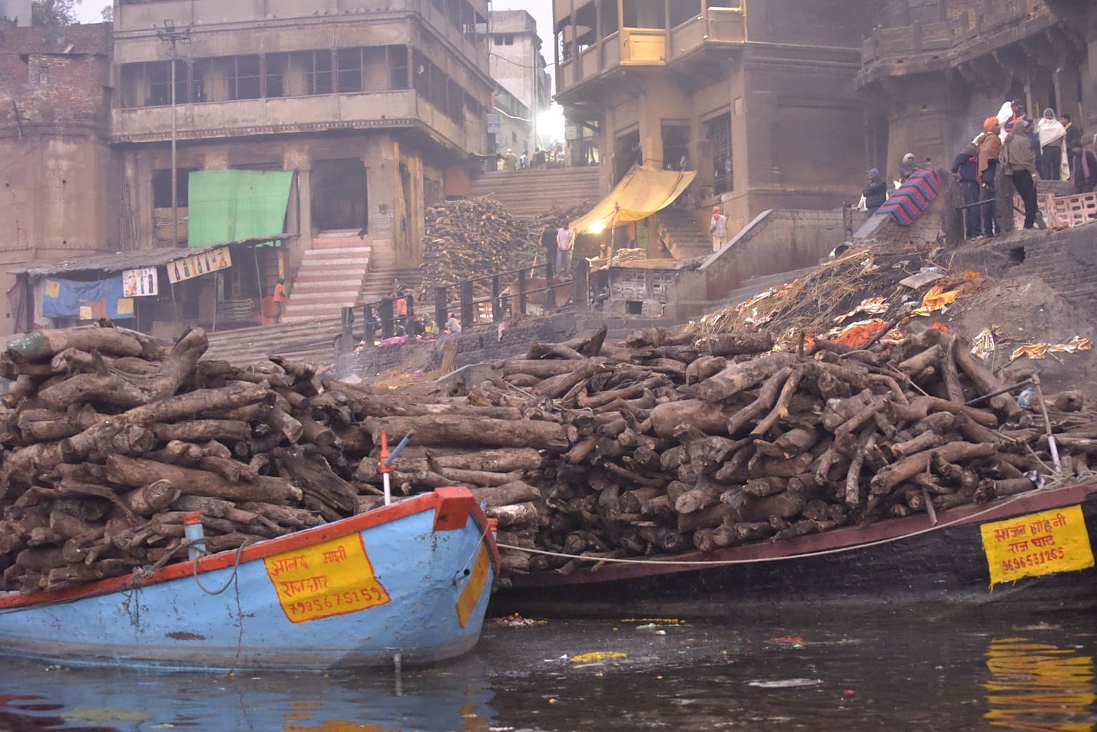 Massed stacks of cremation wood line the ghats behind two small boats on the Ganges in Varanasi, 2019