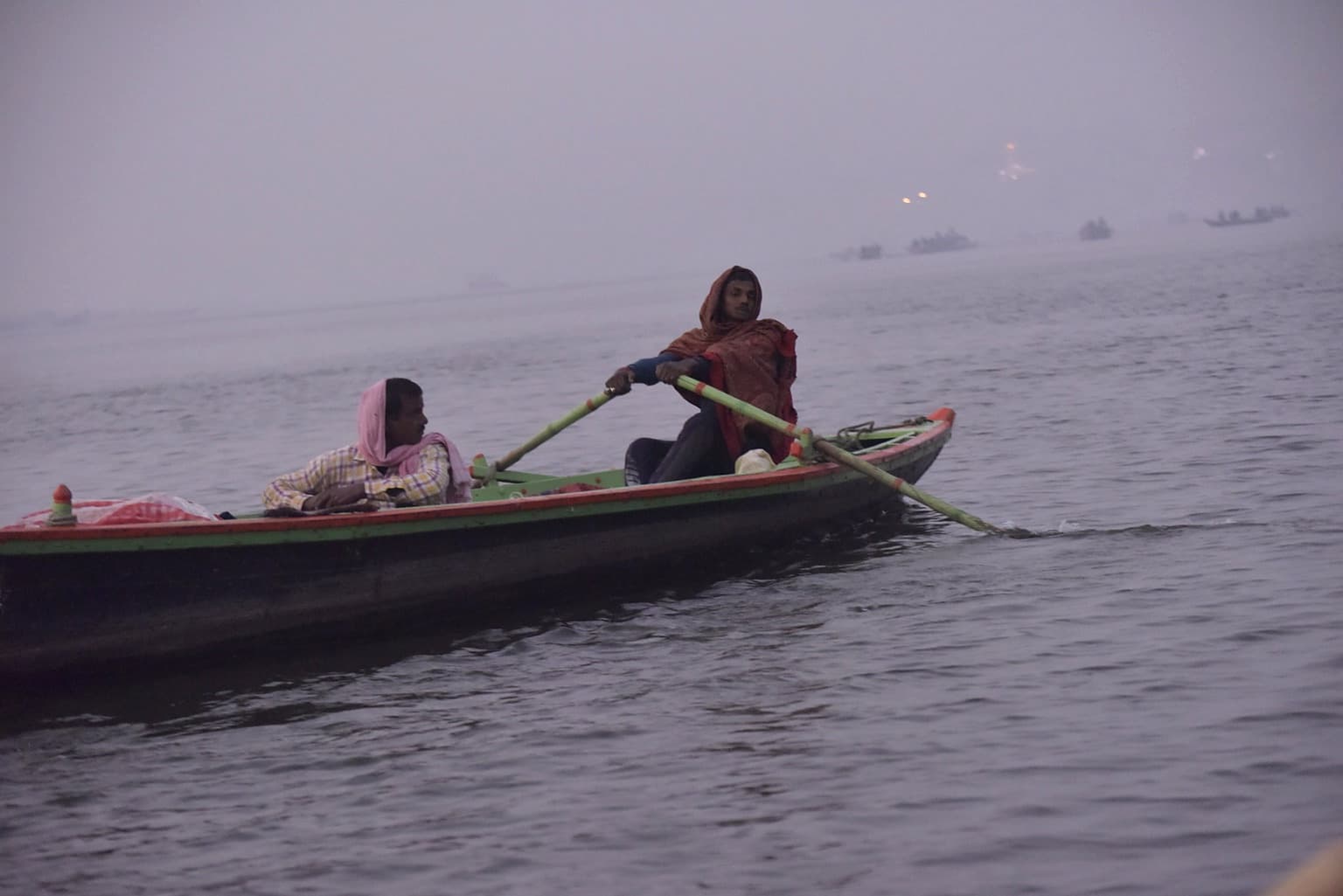 A woman rows a small green and red boat through the haze on the Ganges in Varanasi, 2019