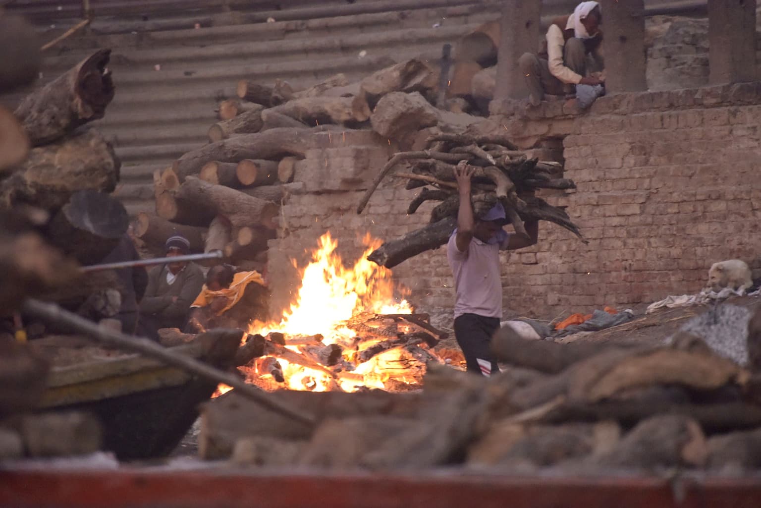 Men work beside an open cremation fire and stacked wood on the ghats of Varanasi, 2019