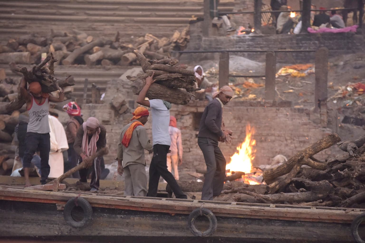 People stand beside a cremation fire among extensive stacks of wood, with a boat crossing the foreground in Varanasi, 2019