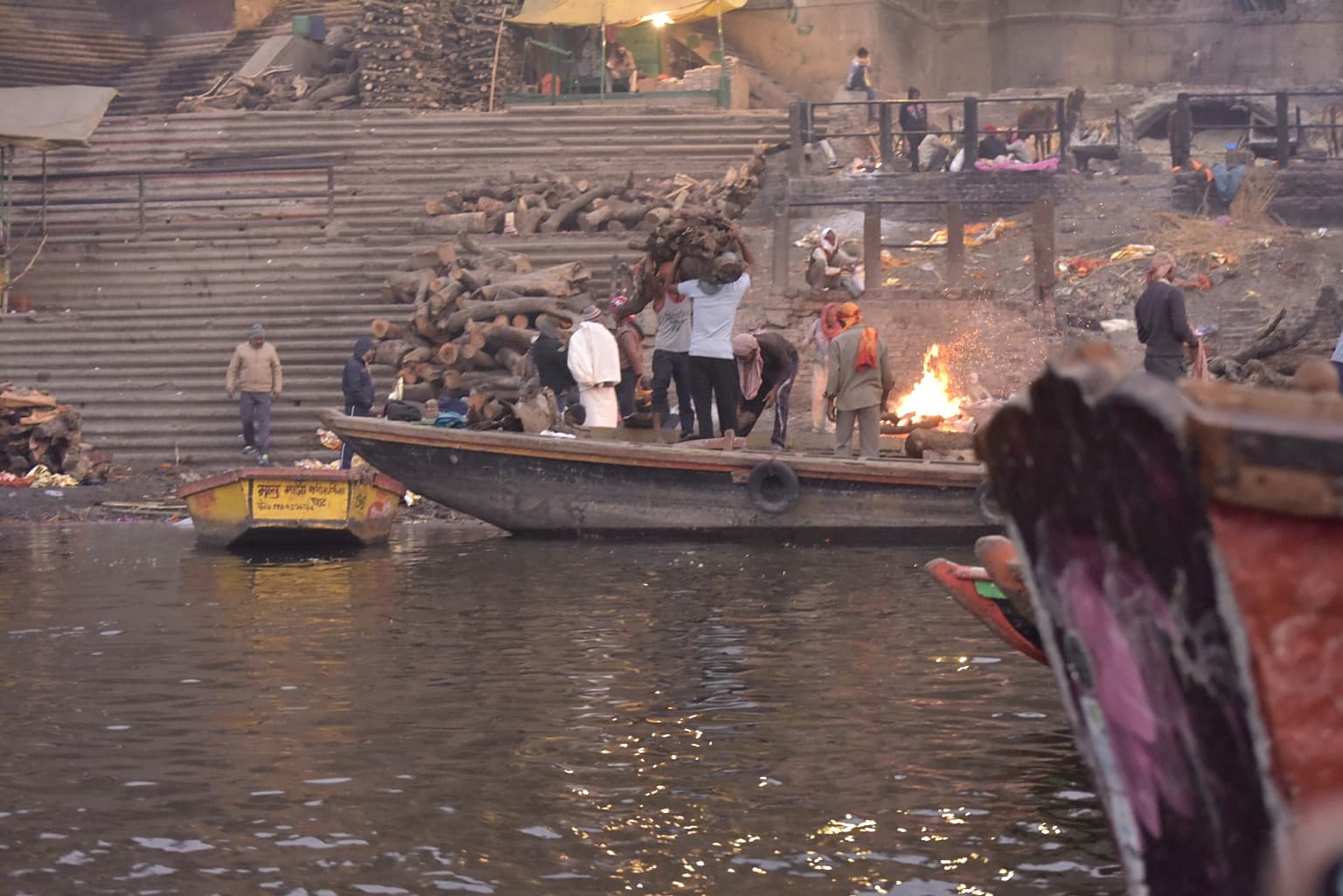 A broad river view shows an active cremation fire, people and layered ghat steps in Varanasi, 2019