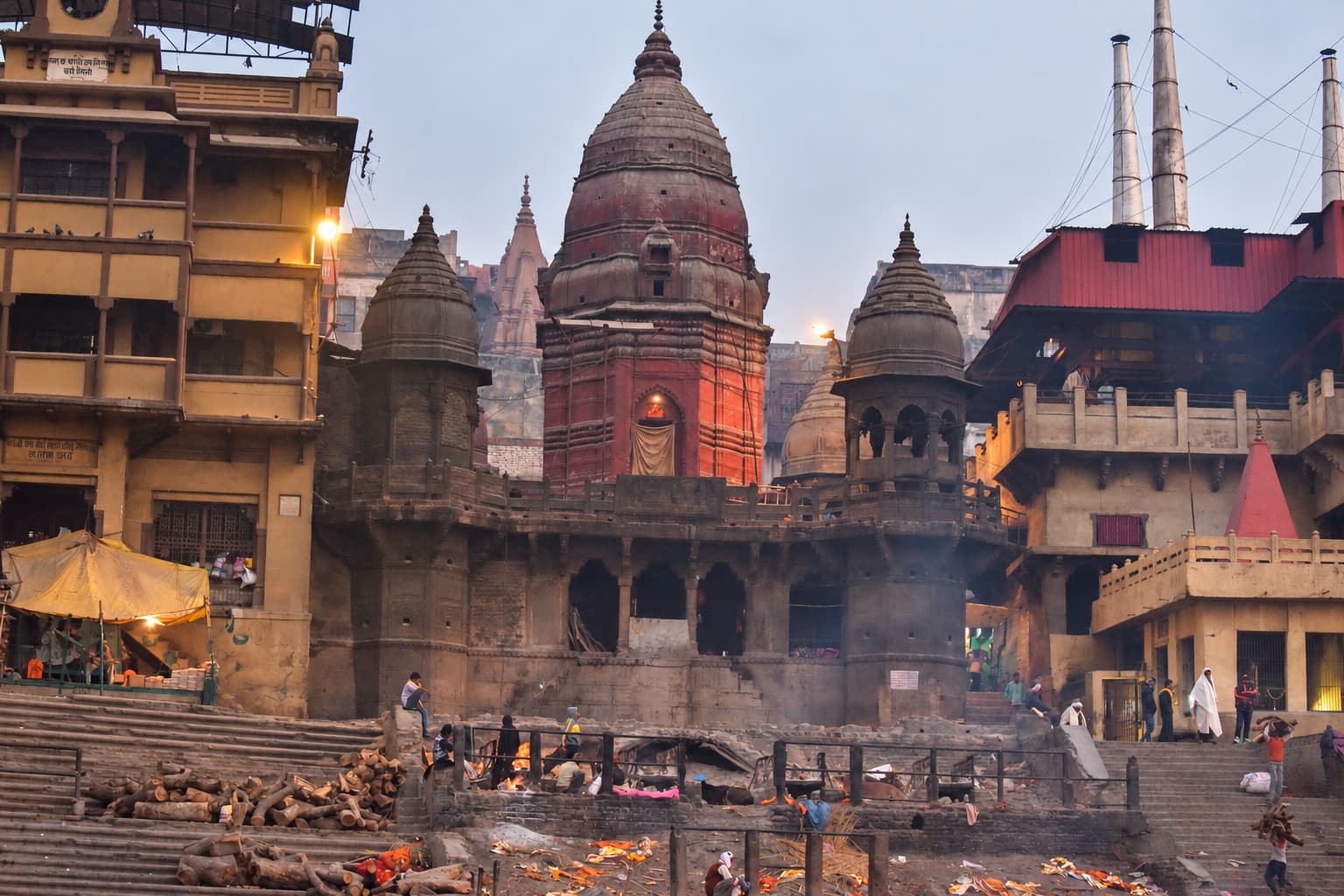 Riverfront temples rise through the morning haze above cremation wood, small fires and people on the ghats of Varanasi, 2019