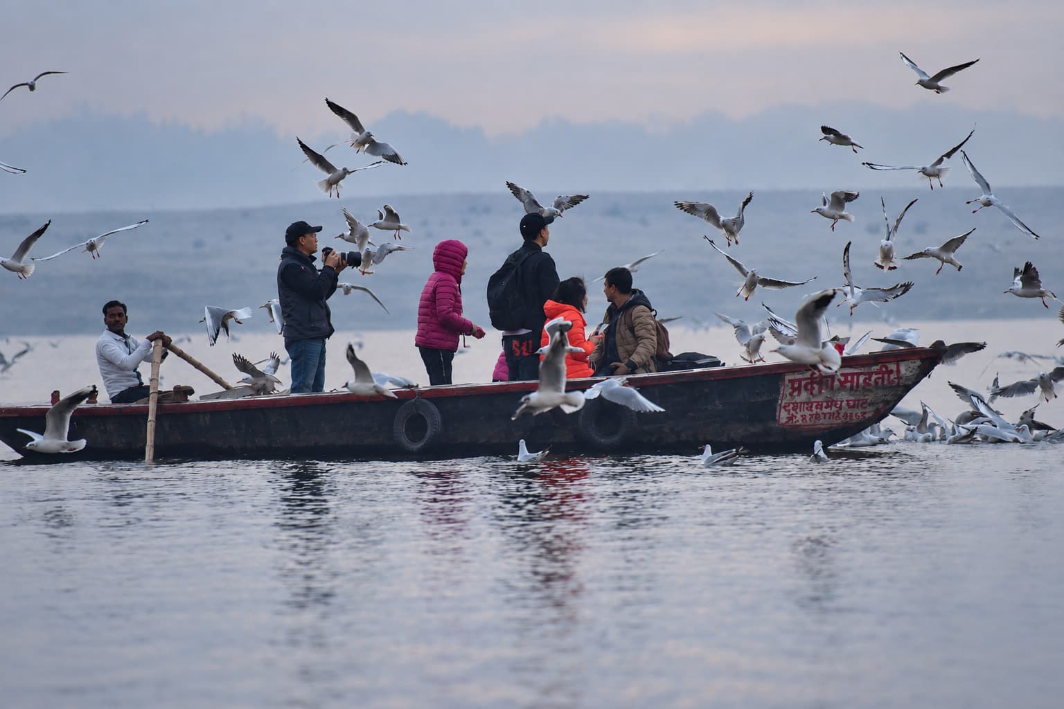 A passenger boat crosses the pale Ganges as birds spread across the morning sky and water in Varanasi, 2019