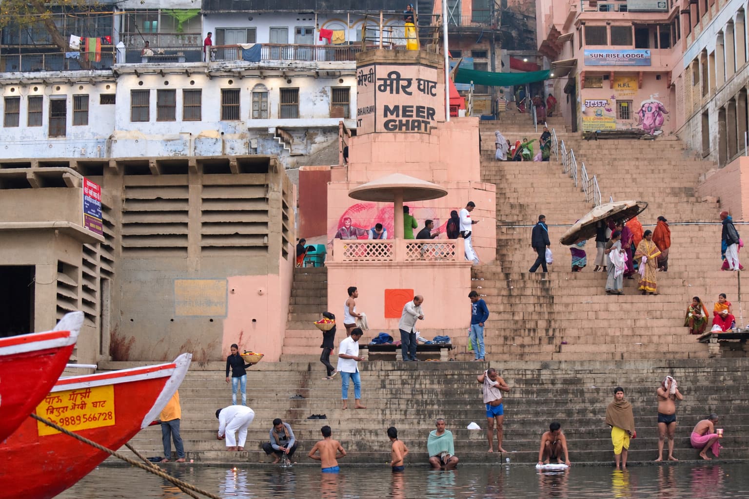 Bathers, vendors and pedestrians occupy the broad ghat steps while a red boat enters the foreground on the Ganges in Varanasi, 2019