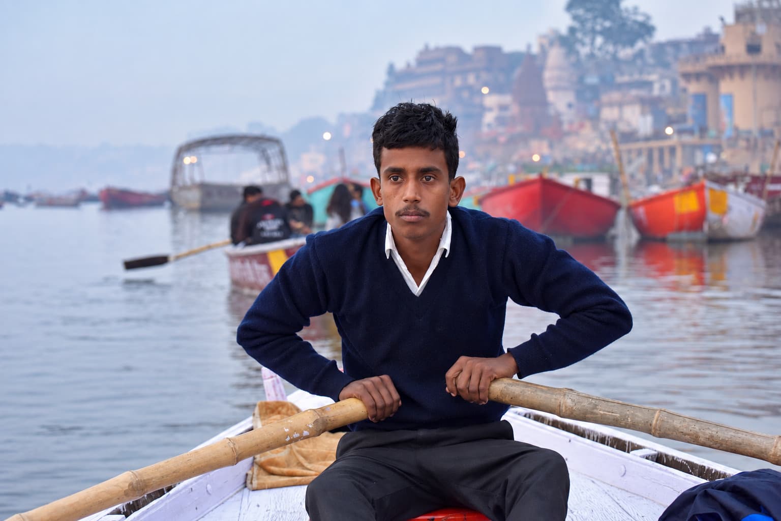 A boatman looks directly toward the camera from his boat on the Ganges, with other boats and hazy ghats behind him, Varanasi, 2019
