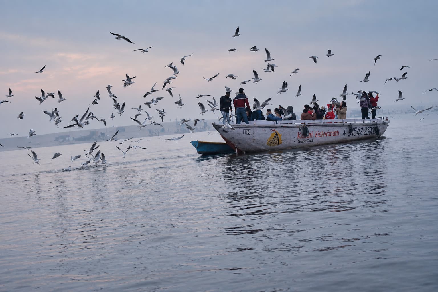 A populated boat moves across the Ganges beneath a broad cloud of birds in the morning haze of Varanasi, 2019