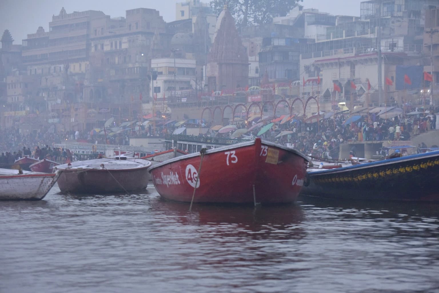 A red passenger boat sits before densely occupied ghats and riverfront buildings in the morning haze of Varanasi, 2019