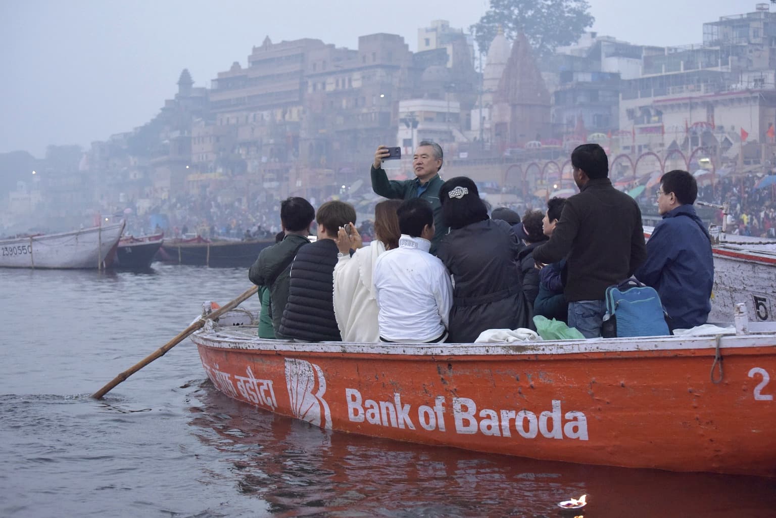 Passengers fill an orange boat on the Ganges while the crowded ghats rise behind them in Varanasi, 2019
