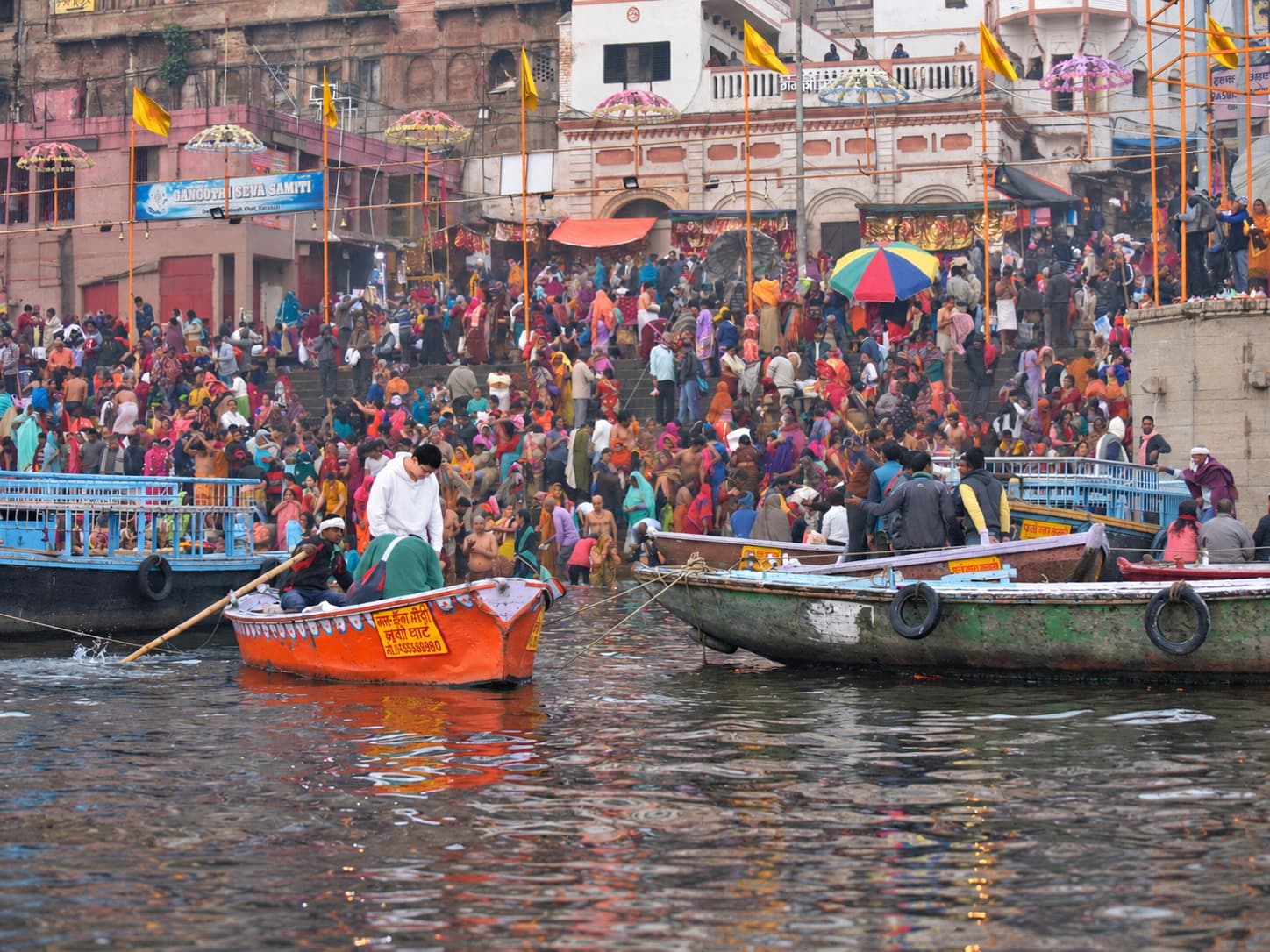 Crowded riverfront steps rise behind working boats, bathers and a brightly painted orange boat on the Ganges in Varanasi, 2019